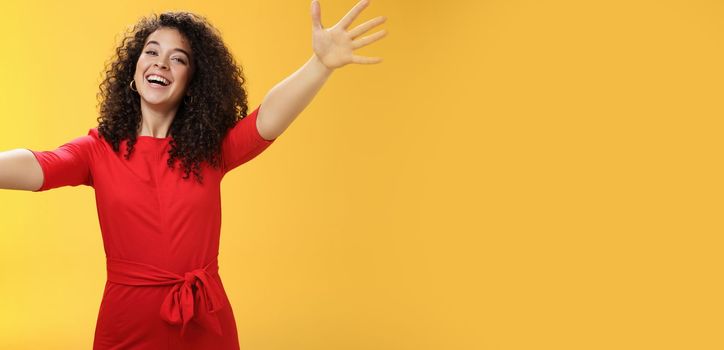 Come into my arms. Portrait of friendly and loving, caring charming woman with curly hair in red casual dress spread hands as wanting give hug smiling broadly at camera giving warm welcome or cuddle.