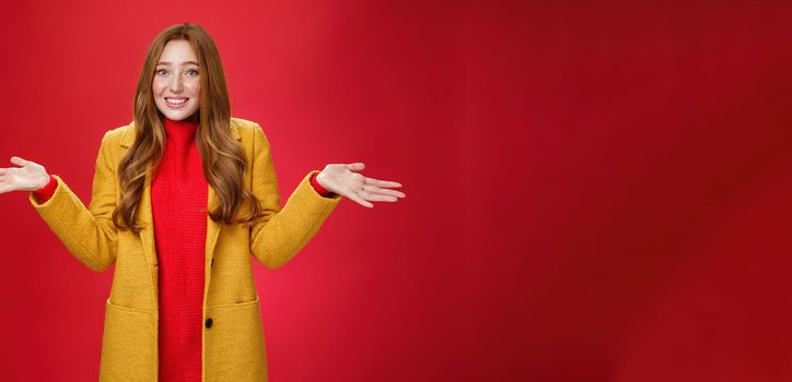 Redhead girl apologizes for being late smiling silly and guilty. with sorry look shrugging as raising hands sideways wearing stylish yellow coat over dress posing against red background.