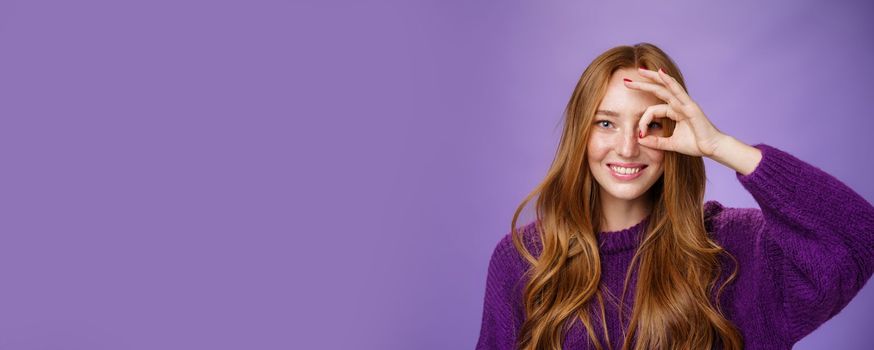 Playful and carefree charismatic redhead female with freckles and long hair showing okay or zero gesture over eye as if peeking joyfully at camera through telescope smiling over purple wall. Emotions and people lifestyle concept