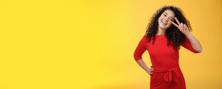 Waist-up shot of friendly tender and feminine cute woman with curly hairstyle tilting head and smiling, laughing and showing victory or peace gesture at camera over yellow background.