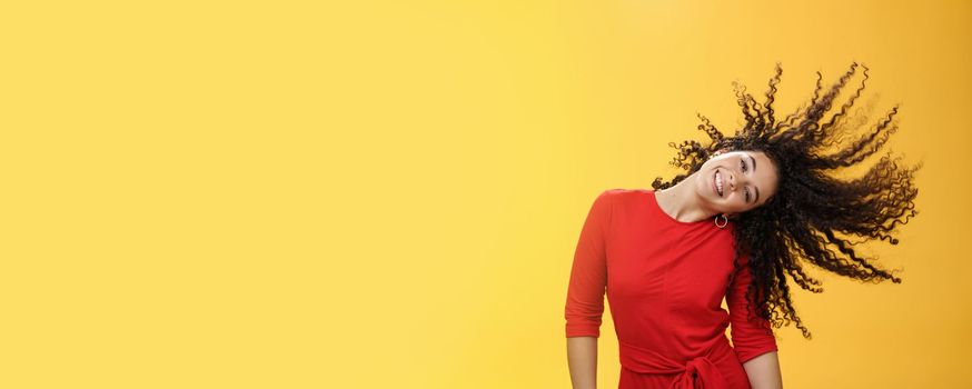 Girl getting carefree and wild waving hair and making sun with curls flying in air smiling broadly tilting head and having fun, dancing delighted and amused feeling playful against yellow background.