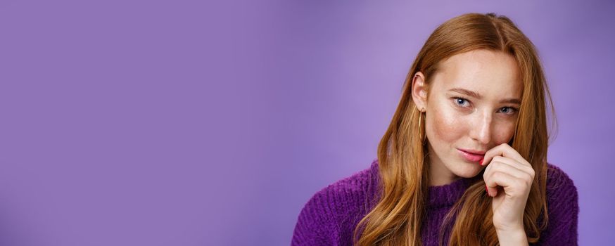 Ginger girl charmes with sensual romantic gaze. Portrait of confident flirting and attractive redhead female tilting head touching lips and looking daring at camera against purple background.