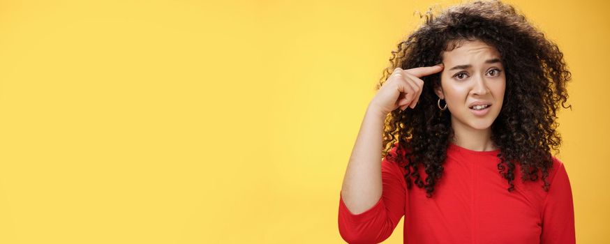 Are you crazy. Portrait of frustrated and confused woman with curly hair holding index finger near temple and raising eyebrows shocked being annoyed with stupid actions of weirdo over yellow wall.