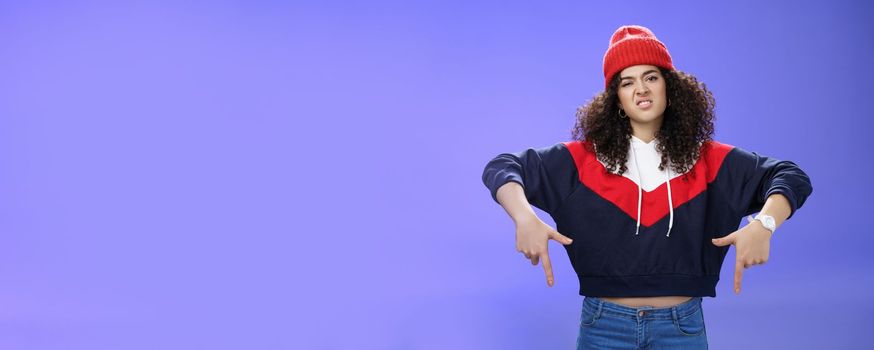 Lifestyle. Snobbish and displeased stylish and cool street girl with curly hair in beanie and sweatshirt pointing down grimacing as feeling unsatisfied and disappointed posing against blue background.