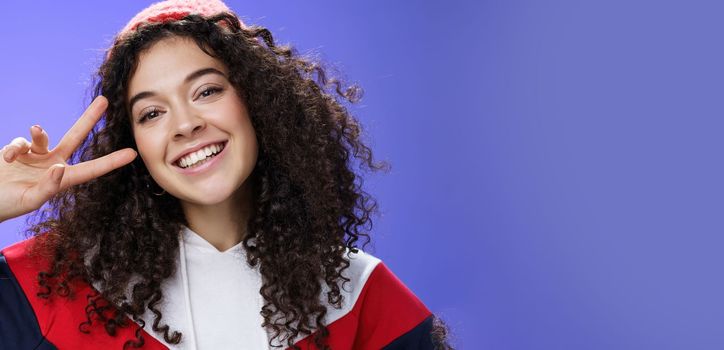 Close-up shot of friendly charming curly-haired woman with warm beanie tilting head joyfully smiling broadly showing victory or peace gesture near face, feeling happy and delighted over blue wall.