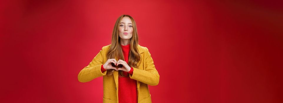 Love you all. Portrait of romantic and stylish good-looking flirty redhead female with freckles and blue eyes folding lips to give kiss showing heart gesture, confessing in sympathy over red wall.