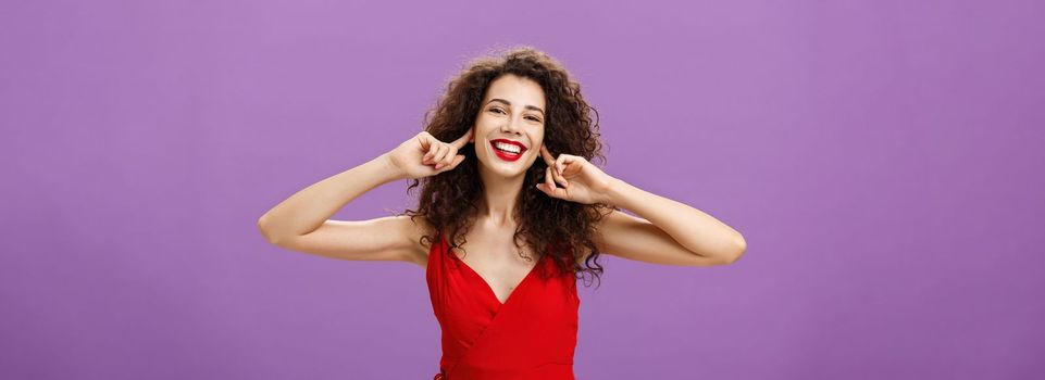 Prefer not listen and live life carefree. Joyful happy outgoing woman in stylish red evening dress closing ears with index fingers not hear about troubles posing delighted and joyful with broad smile.