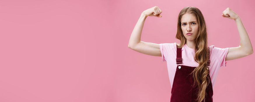 Girl shows she strong and independent. Serious-looking confident slender female raising hands to show muscles feeling self-assured in own power and female strengths posing against pink wall. Copy space