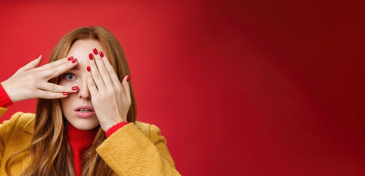 Lifestyle. Stunned and shocked redhead woman cannot believe what terrible thing she sees covering face with palms open mouth from shook and peeking through fingers anxious and scared over red background.