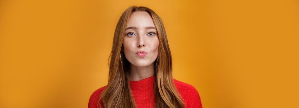 Close-up shot of flirty good-looking sensual redhead female in red knitted clothes folding lips in kiss and gazing at camera as giving mwah feleing romantic and pretty against orange background. Body language, fashion and people concept