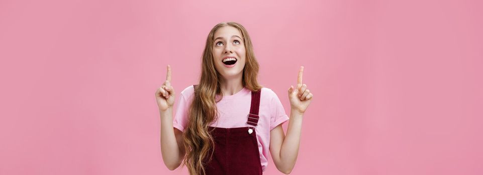 Lifestyle. Amazed and delighted dreamy young teenage girl in stylish corduroy overalls smiling broadly with thrilled smile looking and pointing up joyfully enjoying watching plane flying in sky over pink wall.