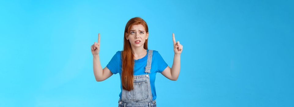 Uneasy worried cute redhead female coworker feel unsure insecure, grimacing displeased intense worried expression, look pointing up, top copy space, have hesitations, stand blue background.