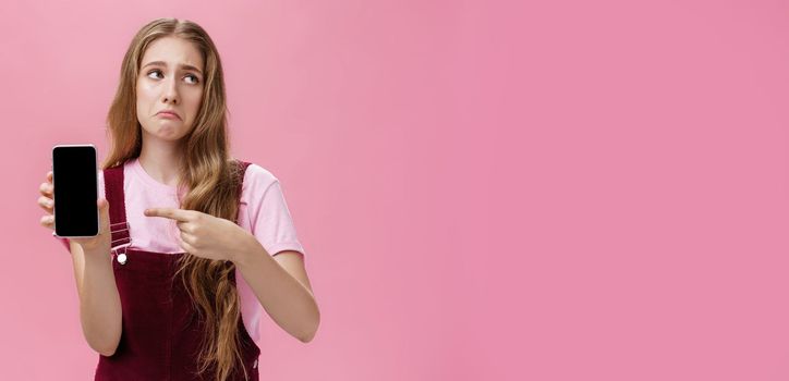 Indoor shot of gloomy displeased and disappointed cute young girl holding smartphone pointing at cellphone screen making upset grimace as if being bothered and dissatisfied against pink wall. Emotions and technology concept