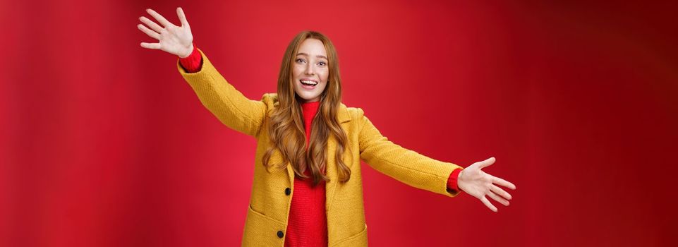 Hey come and hug me friend. Charming playful and emotional redhead female extanding palms forward to cuddle and greet, giving warm welcome smiling broadly posing in yellow coat over red wall.