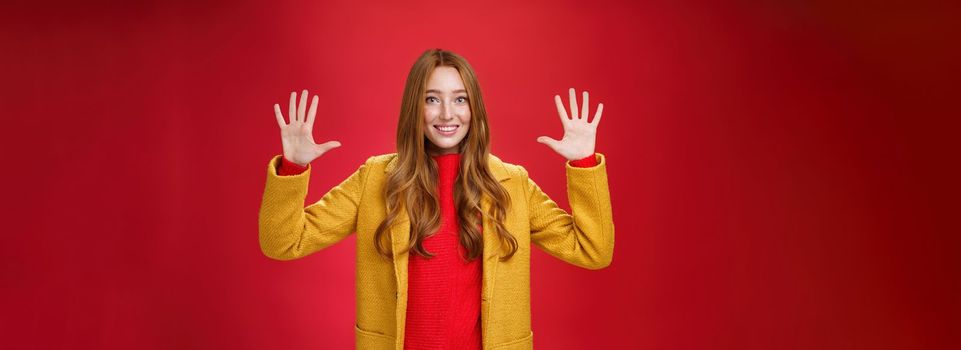 Lifestyle. Cute and tender good-looking friendly redhead woman with blue eyes and freckles in stylish yellow coat showing number ten or ordering dozen of things as smiling happily at camera over red background.