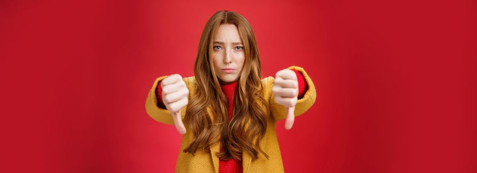 Lifestyle. Portrait of insecure and unconfident cute redhead female reacting to uncool thing showing thumbs down with worried upset expression from dislike and disappointment over red background.