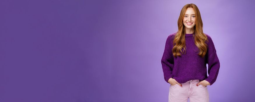 Optimistic attractive ginger girl in 20s wearing purple sweater standing ready and energized over violer background smiling friendly and confident at camera, showing readiness to have fun.