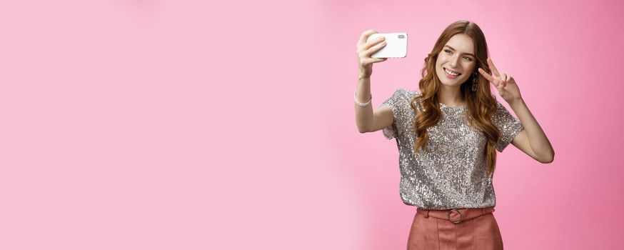 Sociable attractive glamour young caucasian woman taking selfie show victory peace sign smiling broadly raise smartphone look grinning mobile phone display, video calling friends, pink background.