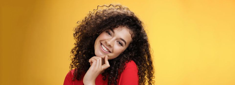 Close-up shot of charming flirty and silly curly-haired young woman making faces as trying get what wants, smiling tilting head on shoulder and touching lip standing cute over yellow background. Romance, people and beauty concept