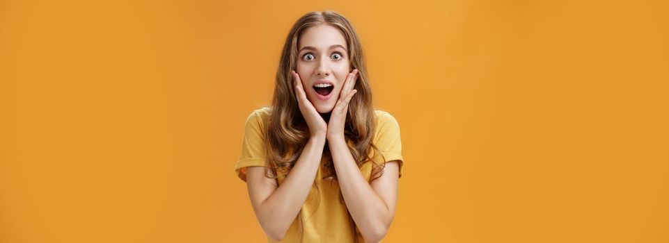 Lifestyle. Waist-up shot of surprised and amused girl learning shocking awesome rumor touching cheeks from amazement opening mouth intrigued gazing at camera focused on interesting story over orange background.