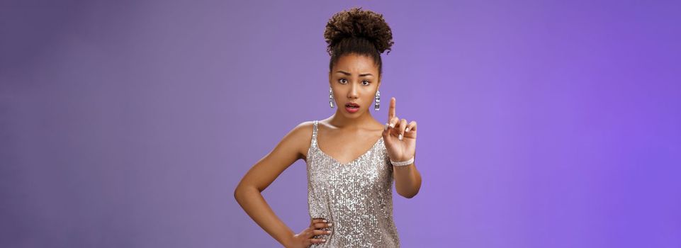 Concerned worried young african-american woman in trendy dress extend index finger nervously look camera shushing prohibiting tell anyone secret frowning afraid, saying taboo, blue background.