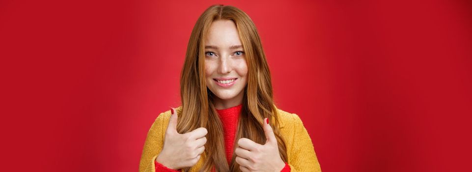 Close-up shot of satisfied happy young 20s woman in yellow coat showing thumbs up gesture in like and approval giving positive feedback enjoying nice event standing over red background, smiling cute.