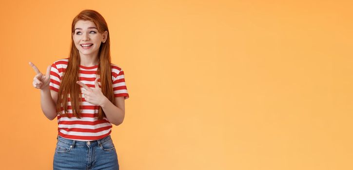 Cute attractive feminine ginger young 20s woman smiling broadly pointing look left happily, reacting amused cool new gadget feature, stand excited upbeat orange background casual urban clothes.
