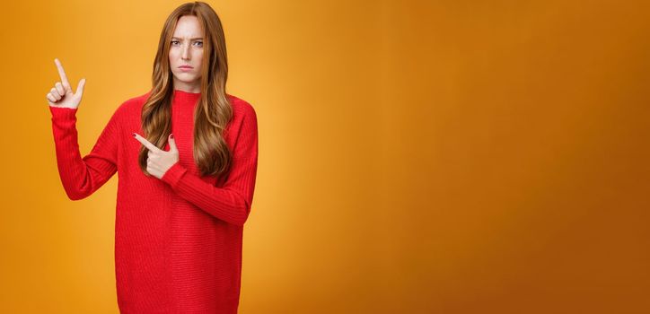 Intense suspicious and doubtful european ginger girl in warm sweater frowning hesitant and confused pointing at upper left corner asking question, looking with disbelief over orange background.