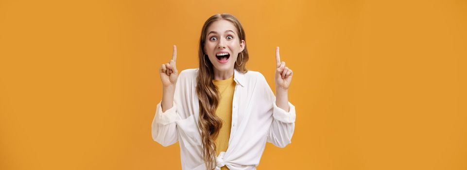 Looking up on bright side. Charismatic happy enthusiastic young attractive woman in white blouse dropping jaw with wide joyful smile pointing up with raised index fingers showing awesome thing.