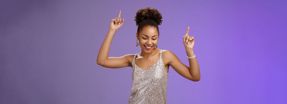 Good-looking carefree african-american girl student have fun prom dancing night club smiling delighted raicing index fingers up joyfully look down, triumphing celebrating good news, blue background.