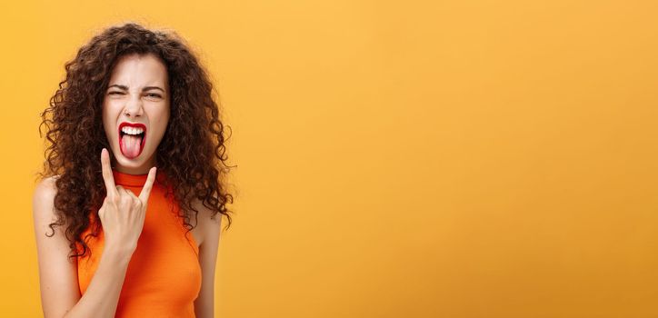 Carefree rebellious stylish urban female with curly hairstyle. sticking out tongue yelling from satisfaction and joy showing rock n roll sign chilling and enjoying cool concert over orange background.