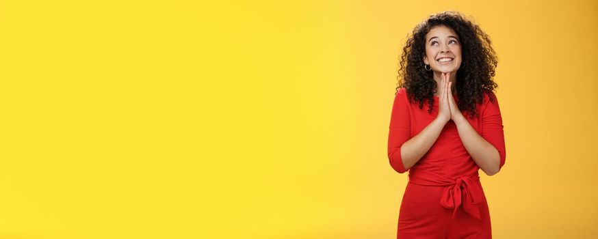 Lifestyle. Dreamy excited cute b-day girl with curly hair in cute red dress rubbing palms together near chest as hands in pray smiling looking up delighted and hopeful making wish over yellow background.