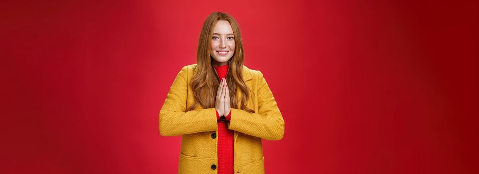 Girl welcomes us with buddhist namaste gesture pressing palms together over chest smiling friendly and delighted with carefree emotions holding hands in pray, posing happily over red background.