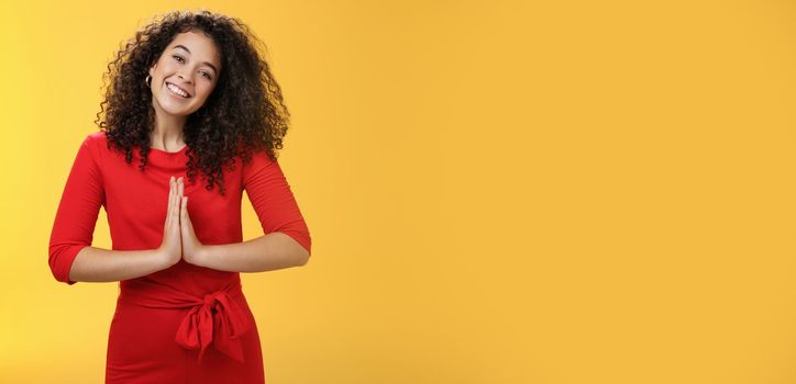 Welcome come inside. Portrait of friendly and polite good-looking female host in red dress with curly hair holding hands in namaste gesture tilting head and smiling as inviting guests in asian style.