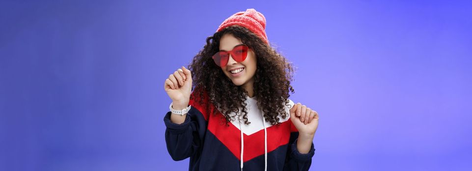 Lifestyle. Stylish and feminine girlfriend dancing having fun feeling upbeat as liking weather feeling warm and cozy in stylish red beanie and sunglasses dancing with raised hands and broad smile over blue wall.