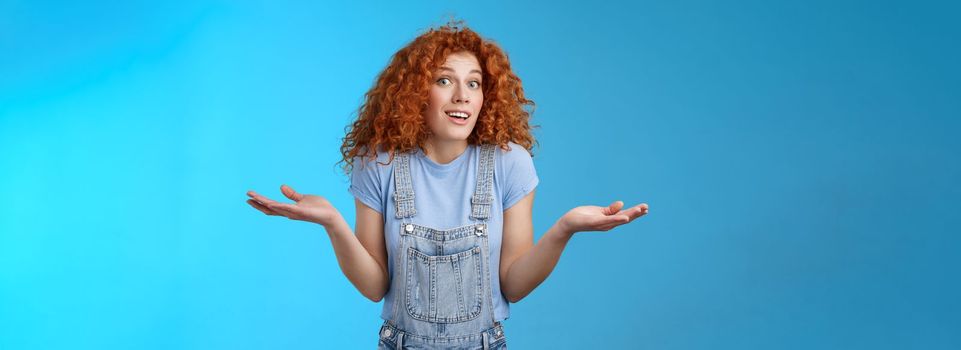 Who knows. Careless clueless attractive playful redhead cute curly-haired girl shrugging unaware hold hands sideways unaware smiling act innocent uninvolved not knwo answer blue background.