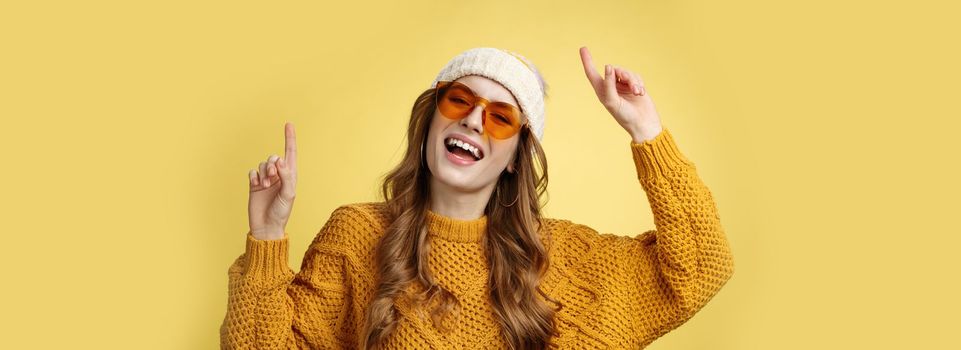 Girl enjoying party have fun smiling broadly happy gazing camera raising index fingers up dancing carefree laughing wearing sunglasses cozy sweater hat, celebrating weekends, yellow background.
