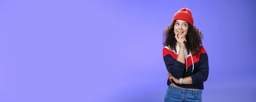 Girl making up idea what snowman built standing cute and silly with thoughtful expression biting finger as thinking, smiling delighted, having interesting thought standing in hat and warm sweatshirt.