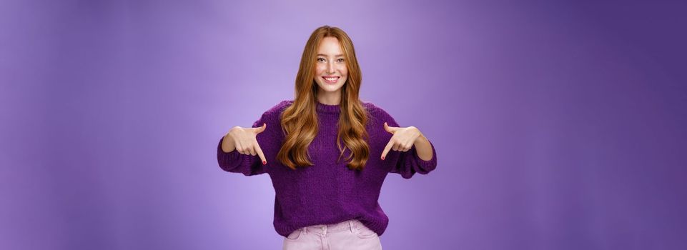 Girl giving recommendation to visit place, pointing down and smiling friendly with delighted and pleasant grin. Portrait of attractive redhead female model in purple warm sweater promoting copy space.
