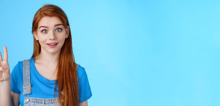 Surprised cute redhead caucasian girl show number three, third, counting fingers, raise eyebrows ambushed, smiling curiously, stand blue background playful tender expression. Copy space