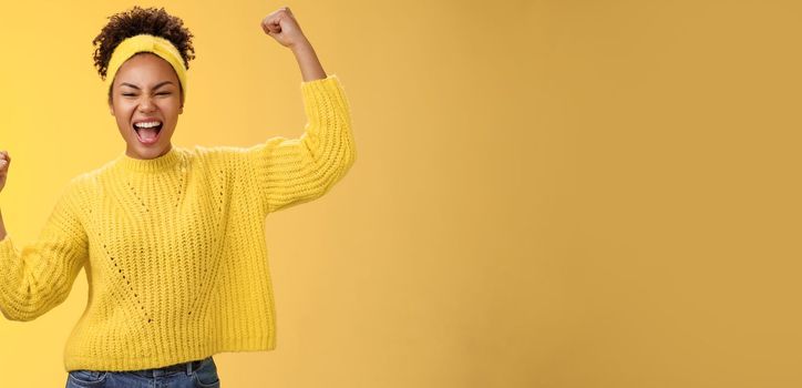 Active outgoing confident cheerful african-american female fan place bet hopefully yelling encourage team win standing raised fists victory celebrating gesture shouting proudly, yellow background.