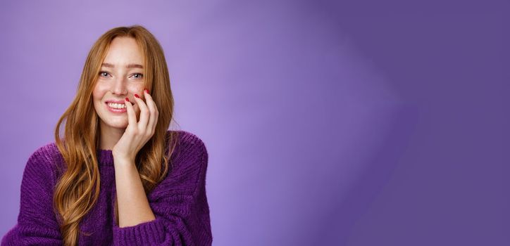 Lifestyle. Close-up shot of feminine and glamour cute redhead girl with freckles in purple knitted sweater holding hand on cheek gently chuckling and smiling broadly at camera over violet background.