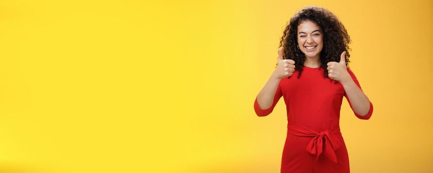 Creative and charismatic happy upbeat woman 25s with curly hair in red dress winking in approval and showing thumbs up with broad smile, satisfied giving positive reply over yellow wall.