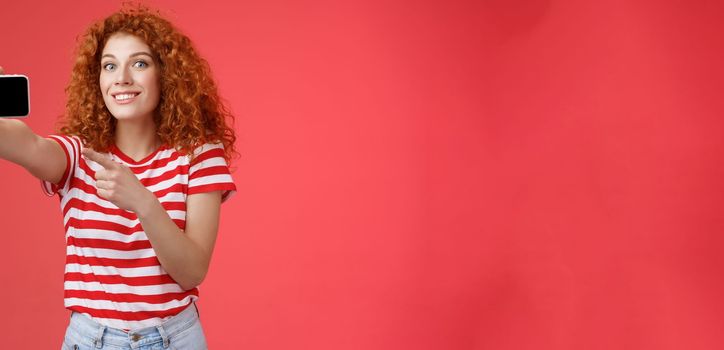 Excited happy good-looking redhead curly female show horizontal smartphone screen pointing display gadget smiling pleased proud beat friends score game standing red background. Technology concept