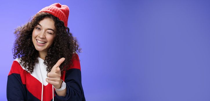 Lifestyle. Friendly-looking sociable and stylish woman with curly hair in red beanie winking joyfully and pointing with finger pistol at camera as greeting mate being cool and confident over blue wall.