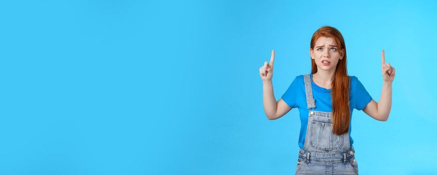 Uneasy worried cute redhead female coworker feel unsure insecure, grimacing displeased intense worried expression, look pointing up, top copy space, have hesitations, stand blue background.