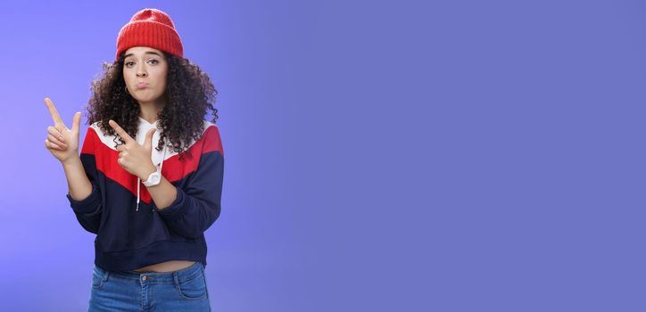 Portrait of silly upset cute curly-haired woman in trendy beanie pursing lips in sadness and frowning making disappointed, regret face pointing at upper left corner unhappy and sad over blue wall.