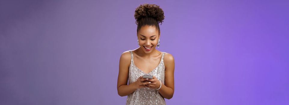 Luxurious good-looking stylish african american woman in silver evening dress holding smartphone chatting texting friends smiling broadly waiting girlfriend near club entrance communicating phone.