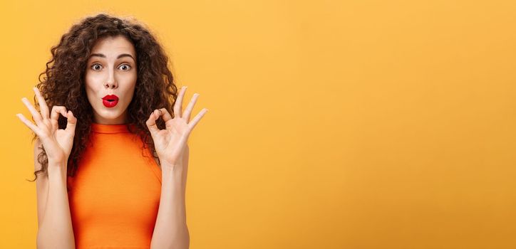 Enthusiastic charming caucasian girl. with curly hairstyle in red lipstick and orange top showing okay gesture being intrigued and delighted folding lips approving and agreeing on awesome adventure.