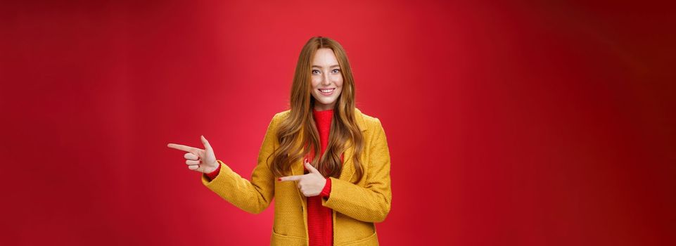 Carefree cute and tender young redhead female student in stylish yellow coat over dress pointing right and smiling with delighted and impressed look showing great place over red background. Advertisement, promotion concept